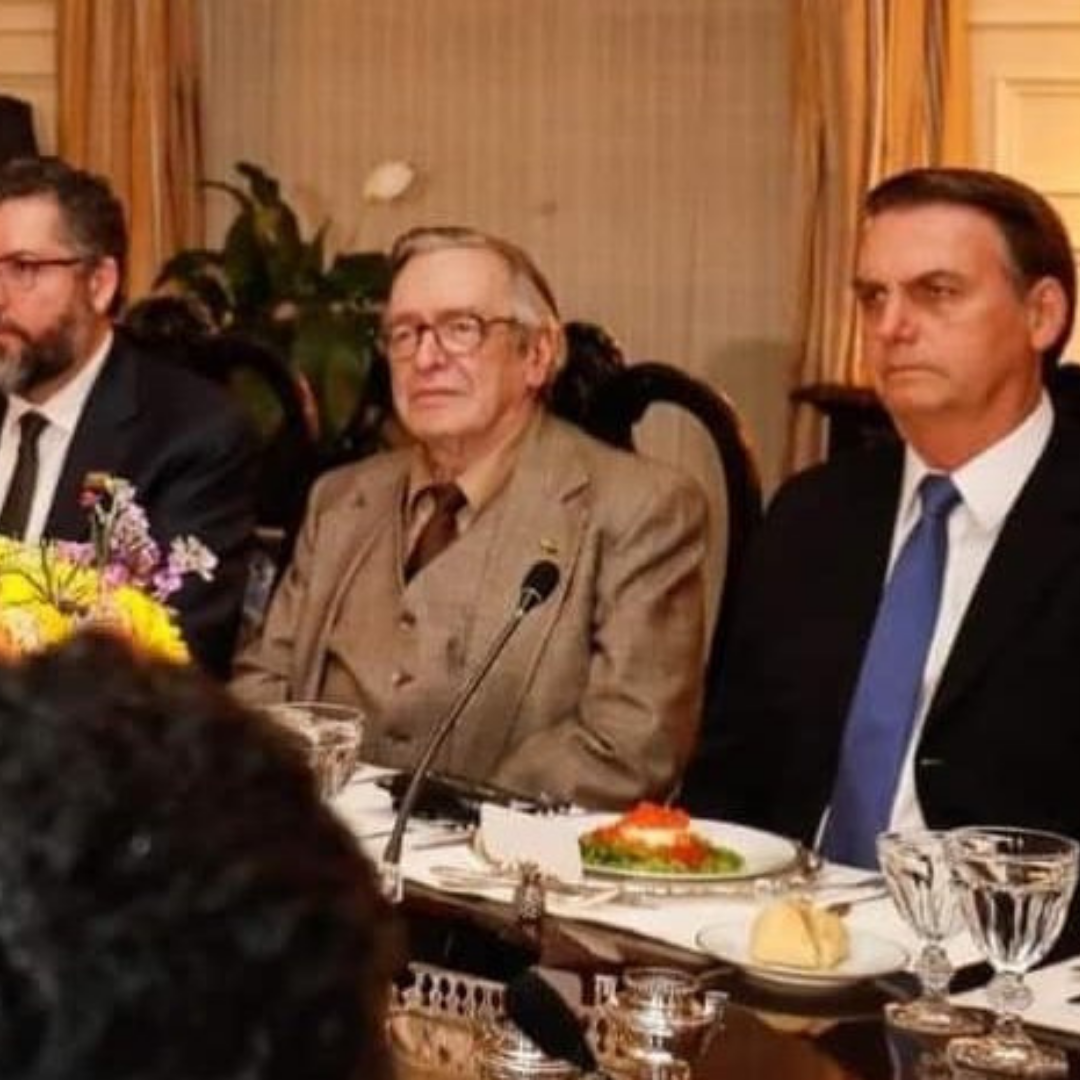 Senhor branco com óculos e terno marrom, Olavo de Carvalho, posa ao lado do presidente Jair Bolsonaro, homem branco de terno preto e gravata azul