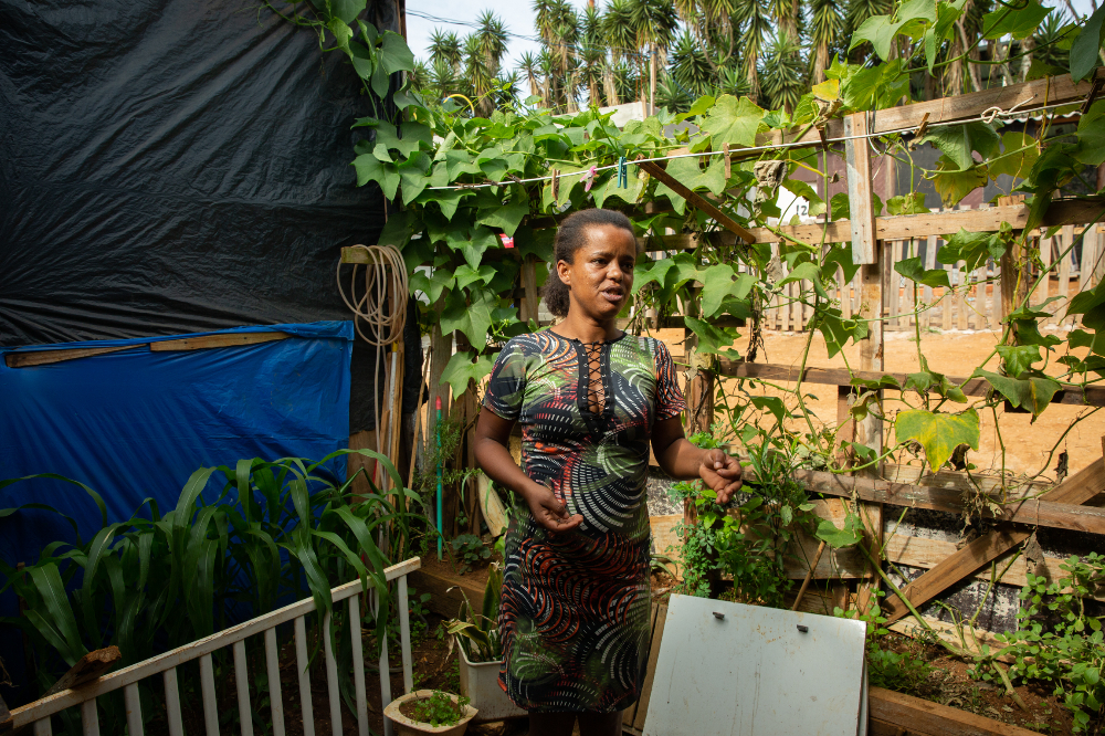 Zenaide é uma mulher negra, ela veste um vestido estampado justo ao corpo; ao seu redor há inúmeras plantas