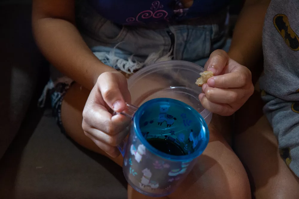 A imagem mostra as mãos de uma mulher segurando uma xícara de café e um pedaço pequeno de bolacha