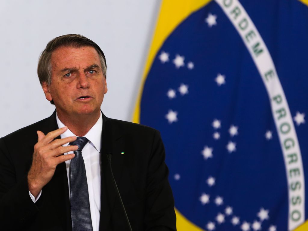 2022 e eleições: Lula vs Bolsonaro: desinformação e risco de golpe segundo pesquisadores 53 Presidente Bolsonaro em frente à uma bandeira do Brasil; ele veste um terno preto com camisa branca