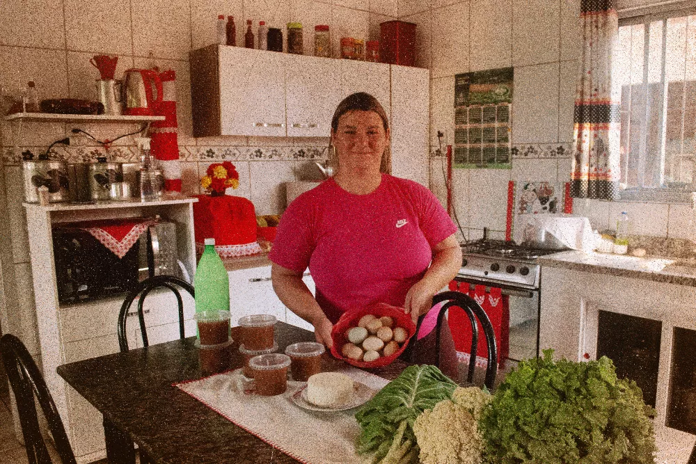 De modelo internacional à extinção: como morre uma política pública 38 Uma mulher branca está em uma cozinha, ela segura um pote com ovos; na mesa à sua frente estão algumas verduras que ela colheu de sua horta