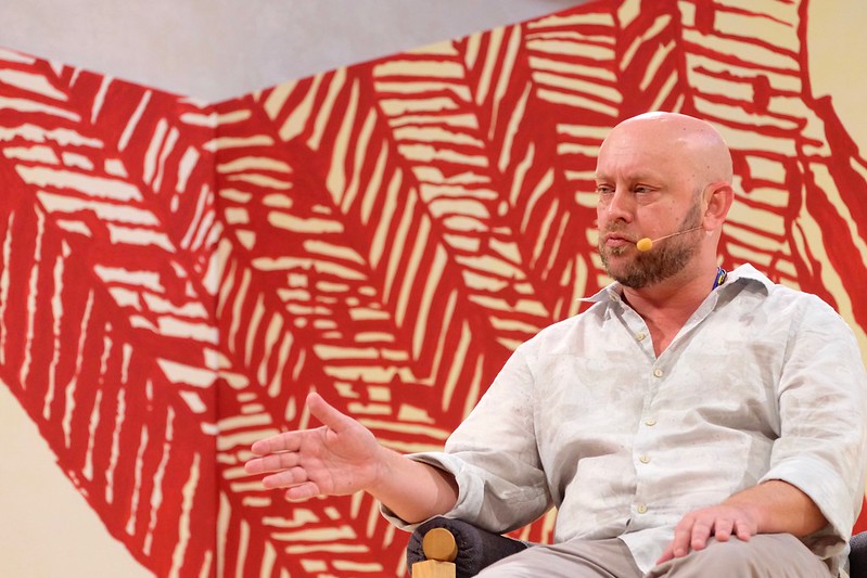 Simas é um homem branco, careca e com barba castanha clara; ele veste um camisa branca; atrás dele há um painel vermelho