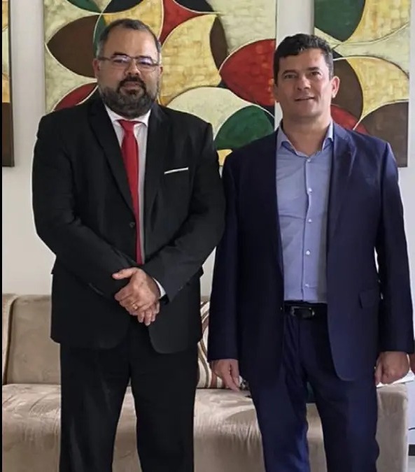 Uziel Santana e Sergio Moro são dois homens brancos com cabelos e olhos castanhos; Uziel tem uma barba média, usa óculos e veste um terno preto, já Moro usa um terno azul