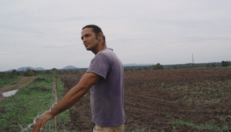 Fernando era um homem pardo com cabelo e olhos castanhos; ele veste camiseta cinza com calça bege; na imagem ele está apoiado em uma cerca, em meio a um campo aberto