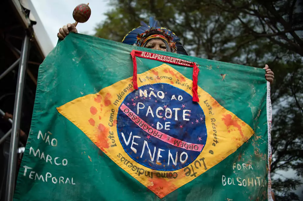 Txai Suruí: “Não estão queimando só a Amazônia, estão queimando as pessoas de lá também” 14 Indígena segura a bandeira do Brasil nas mãos; ela está manchada de sangue e com algumas frases escritas como "não ao pacote do veneno", "Fora Bolsonaro" e "Não ao Marco Temporal"