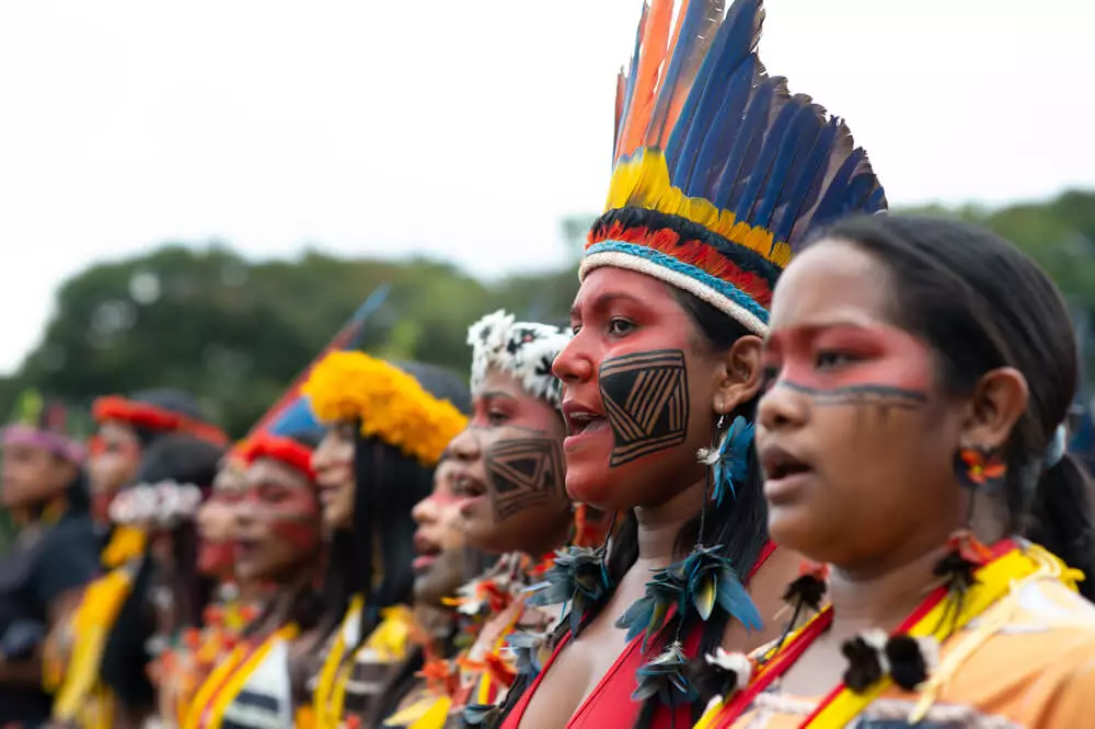 Txai Suruí: “Não estão queimando só a Amazônia, estão queimando as pessoas de lá também” 12 Jovens mulheres indígenas organizadas em fileira durante ato do ATL 2022. Elas vestem cocar e outros adereços tradicionais, além de pintura facial étnica