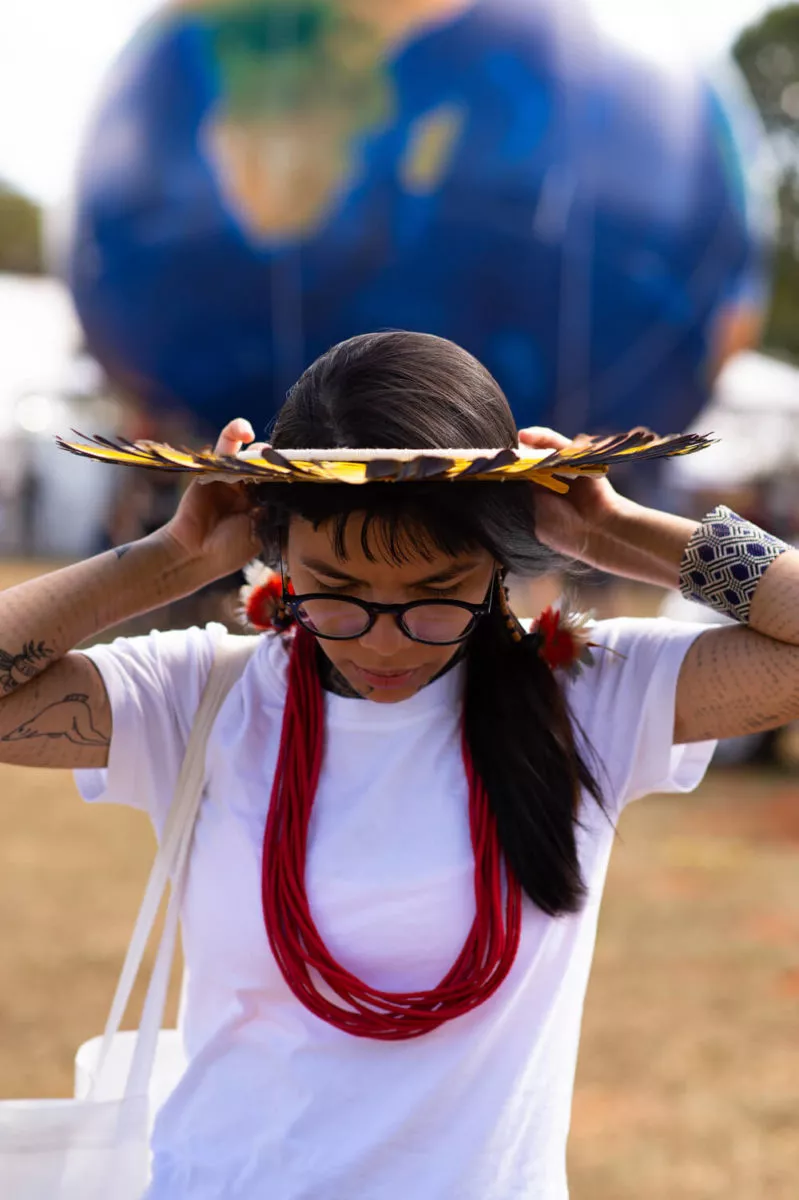 Txai Suruí: “Não estão queimando só a Amazônia, estão queimando as pessoas de lá também” 16 Txai é uma jovem indígena com cabelos castanhos. Ela usa um cocar na cabeça, camiseta branca, colar vermelho e brinco de pena. Na foto Txai esta ajeitando o cocar e olhando para o chão
